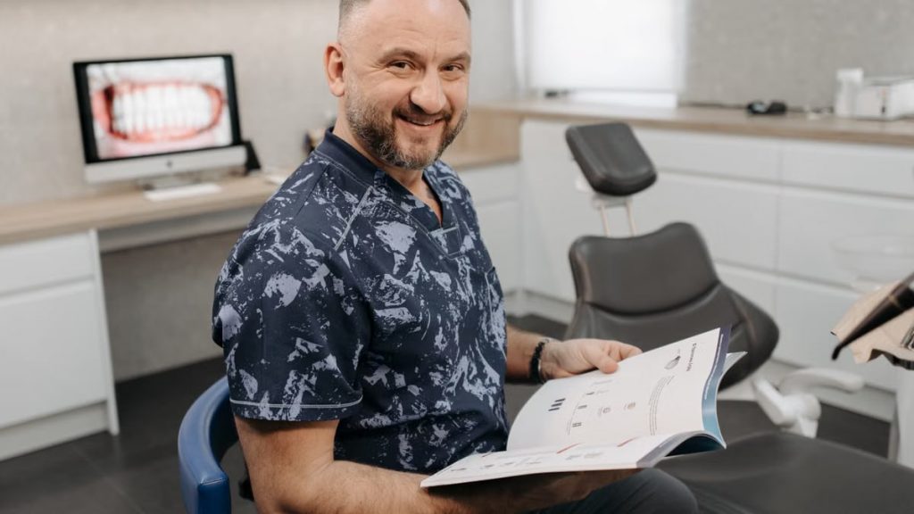 Smiling dentist holding a book, sitting in a dental office with equipment around.